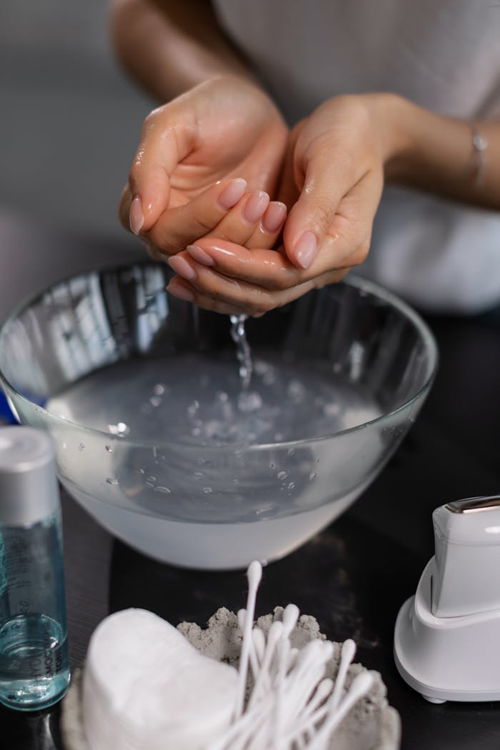 Close-up of hands rinsing in a glass bowl of water, surrounded by skincare items.